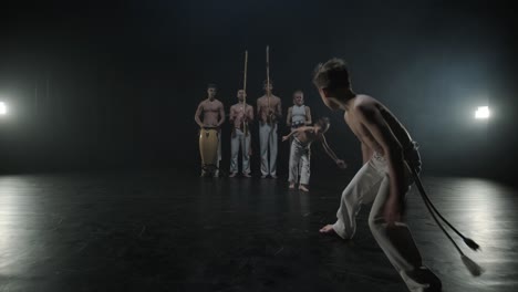 group of a professional dancers are practicing capoeira in darkness against a spotlight on a black background of studio. afro-brazilian martial art that combine elements of dance.