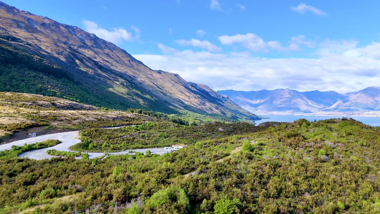 Daytime camera pan reveals winding road, lush hills, and distant lake under clear blue sky