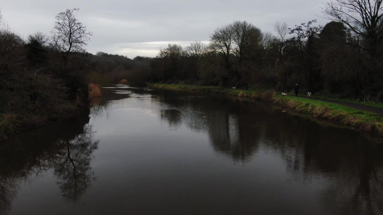 Ripresa con drone di un fiume con persone che portano a spasso i loro cani a destra dell'inquadratura. Cielo mattutino che mostra alcune sfumature di colore e un riflesso luminoso dalla superficie del fiume