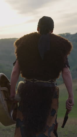 actor in tribal furry clothes walks carrying shamanic drum against old hills backside view. dramatic scene at sunset. siberian ethnic traditions