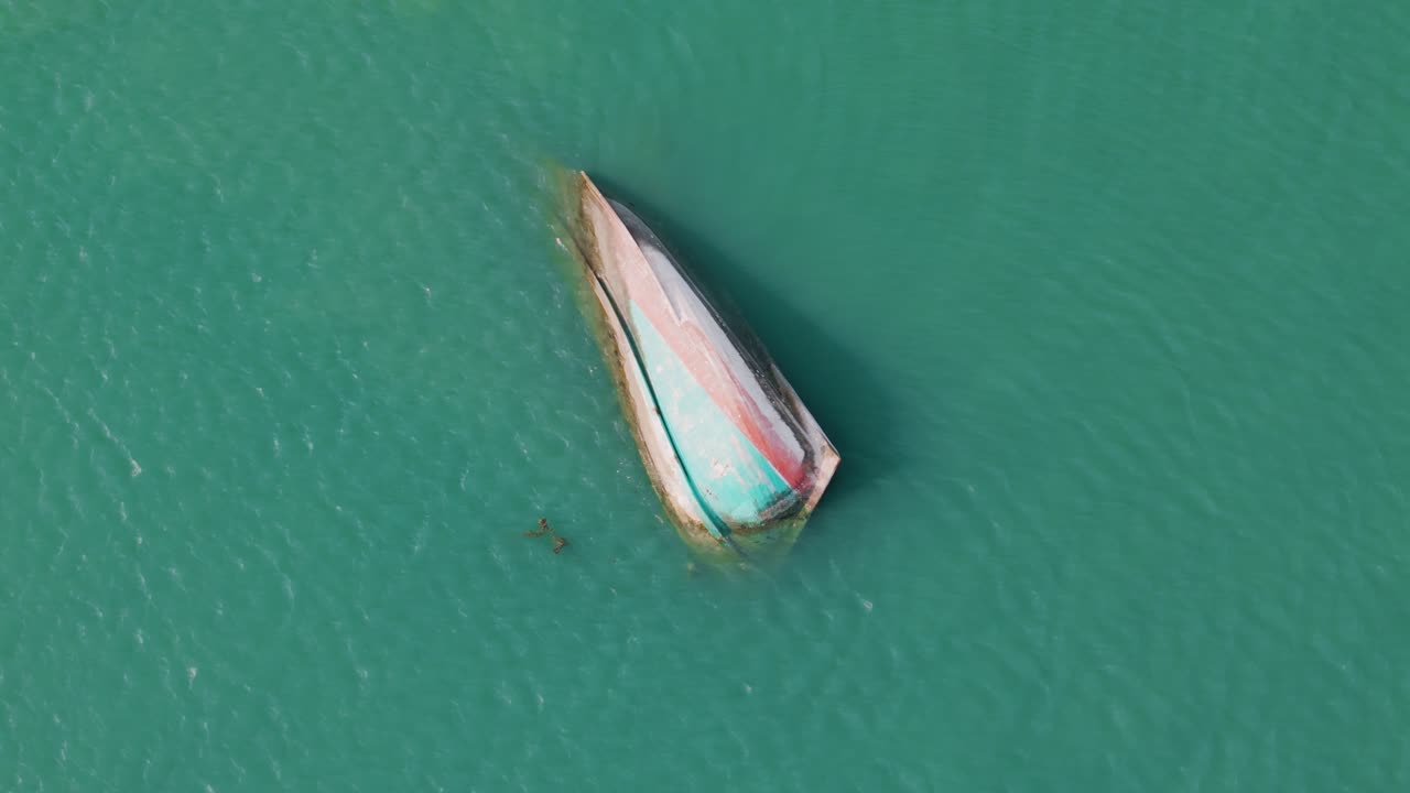 Top-down rotating aerial view of half-sunken colorful fishing boat in crystal clear turquoise tropical bay