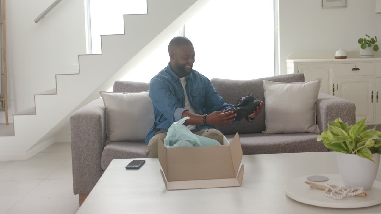 Adult African American male seeing open box on sofa, lifting and checking black shoe quality