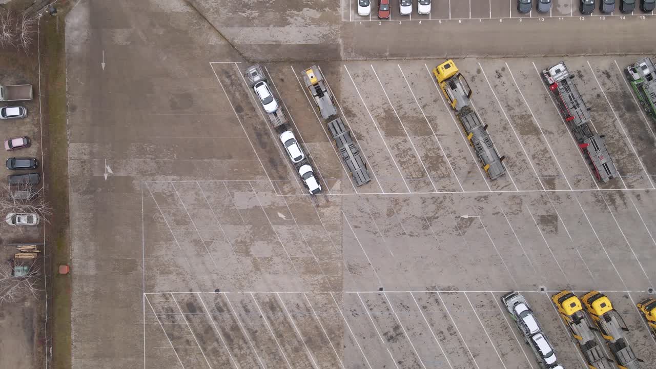An aerial view of a large parking lot featuring car transport trucks loaded with vehicles. The space is mostly empty, with some scattered cars and clearly marked parking spaces.