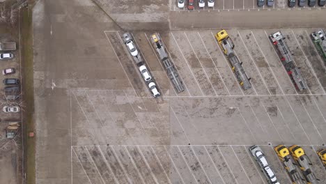 an aerial view of a large parking lot featuring car transport trucks loaded with vehicles. the space is mostly empty, with some scattered cars and clearly marked parking spaces.
