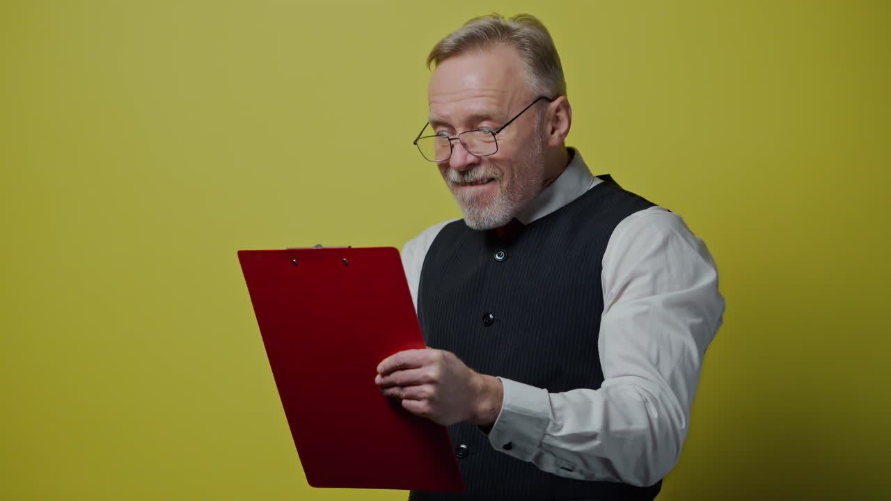 Funny mature man with a folder isolated on yellow wall. Happy old man in glasses writes on paper and depicts funny face. Positive emotions.