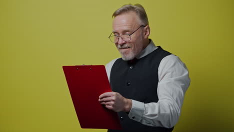 funny mature man with a folder isolated on yellow wall. happy old man in glasses writes on paper and depicts funny face. positive emotions.