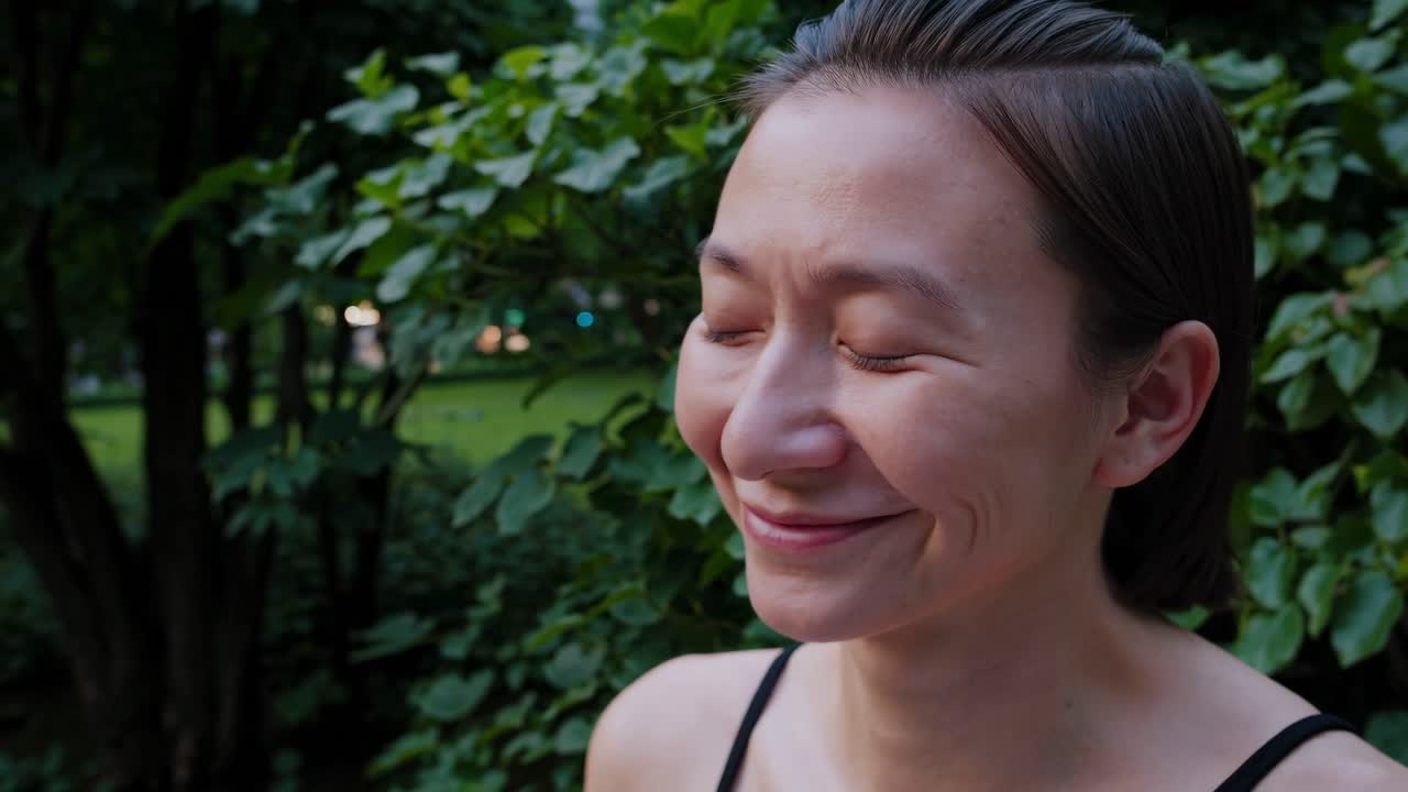 Woman enjoying a serene moment outdoors, eyes closed, surrounded by lush greenery, transitioning from calm reflection to a gentle smile, capturing a peaceful emotional journey