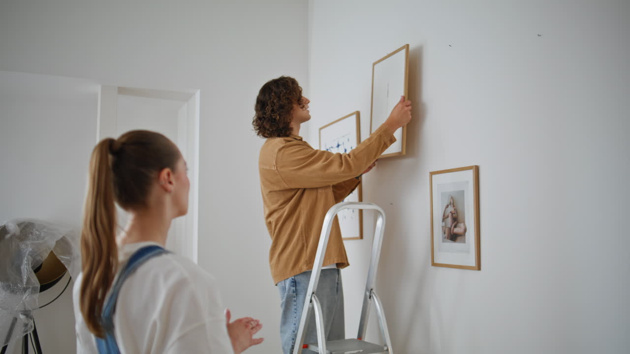 Man ladder hanging picture while woman observing in living room. Family couple