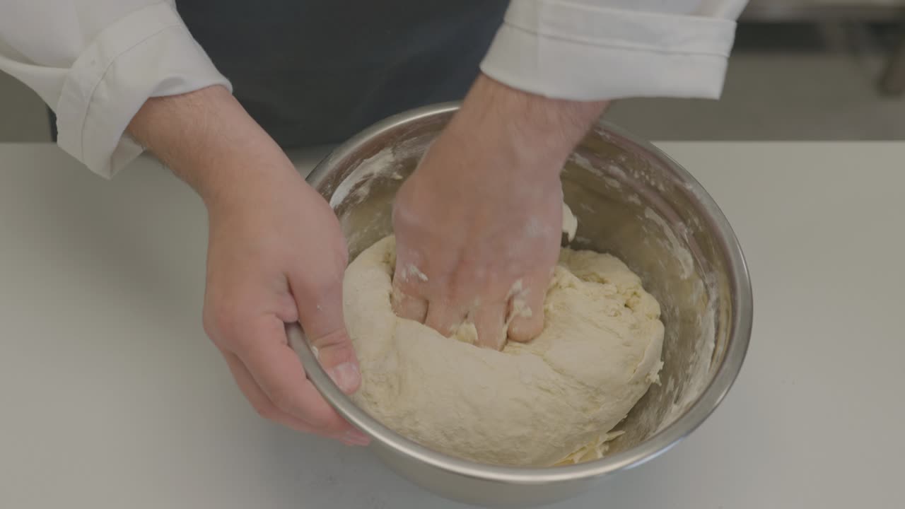 A baker skillfully kneads dough in a large metal bowl