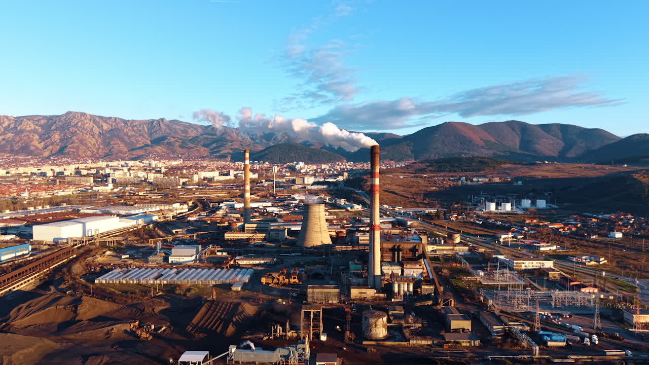 Industrial site with smokestacks and mountains in background during daytime. An industrial area shows buildings, smokestacks, and mountains in clear daylight with city far away