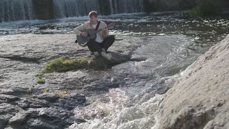 young man playing guitar sitting on the bank of a mountain river on a background of rocks. concept of freedom relaxation. place