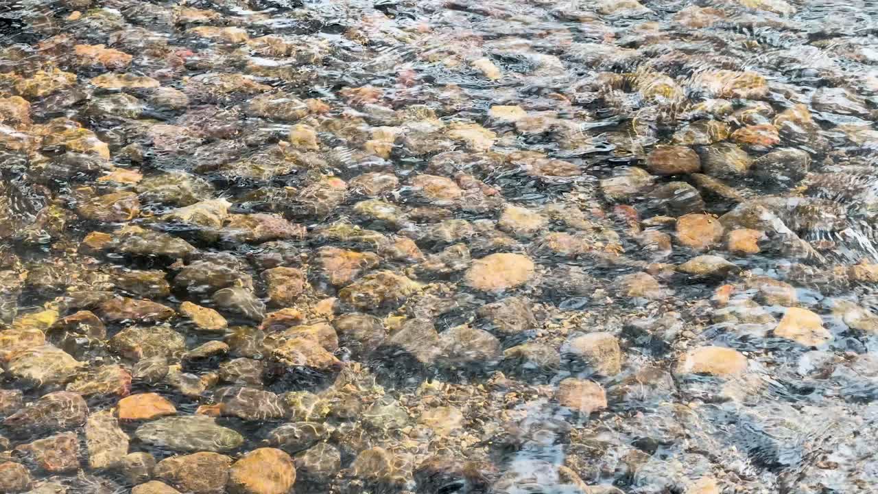 Inquadratura statica dall'alto di acqua trasparente che increspa su ciottoli colorati in un ruscello.