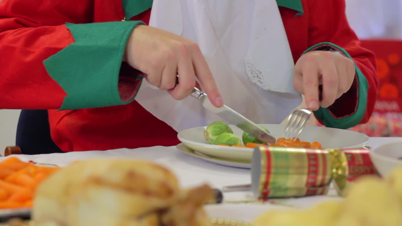 el duende de papá noel comiendo la cena navideña en una mesa llena de comida que incluye pavo y verduras.