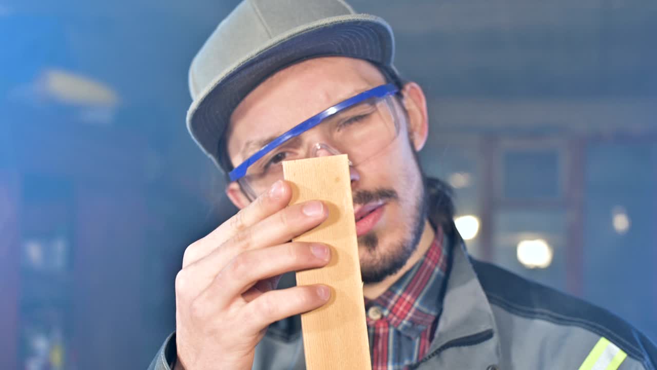 joven carpintero barbudo concentrado con gafas de protección y una gorra comprueba la pieza de trabajo
