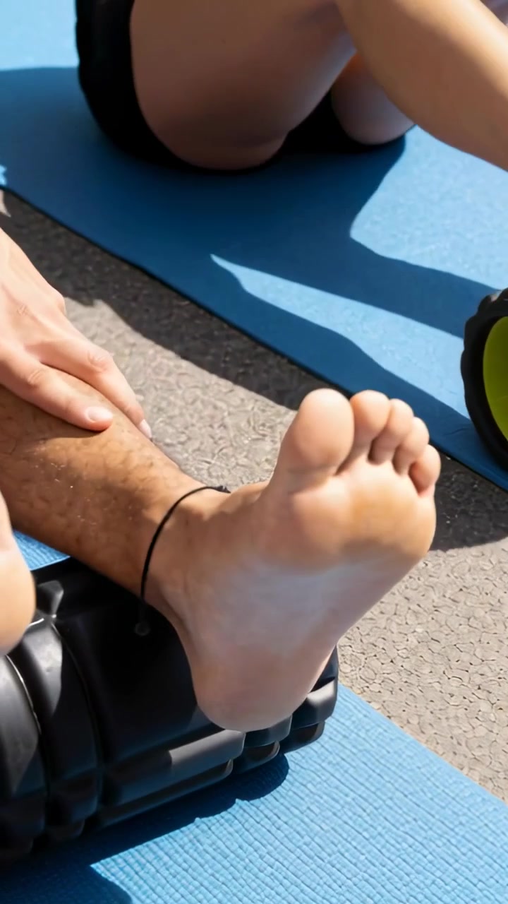 Vertical video: Placing foot on black roller, pal in shorts pressing rolling calf on mats for rehab