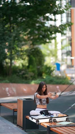 teenage girl using phone on park bench with luggage