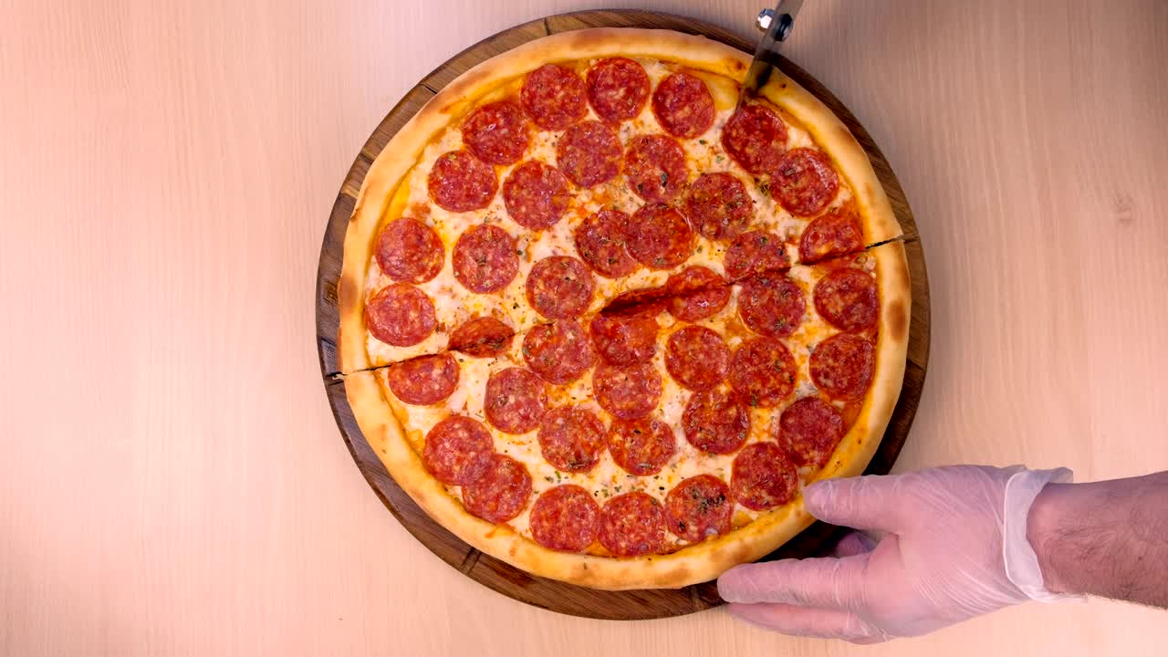 요리사는 살라미 피자를 조각으로 잘라줍니다. 고무 장갑을 입은 손으로 가까운 위쪽을 보세요.