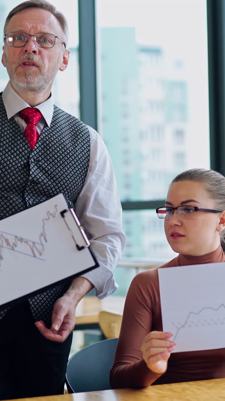 Elderly businessman and a secretary in the office. Attractive company worker sitting at the desk and talking to her boss about some graphics. Vertical video