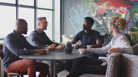 Four-male-creative-colleagues-talking-at-a-table-in-their-workplace-cafe,-three-quarter-length