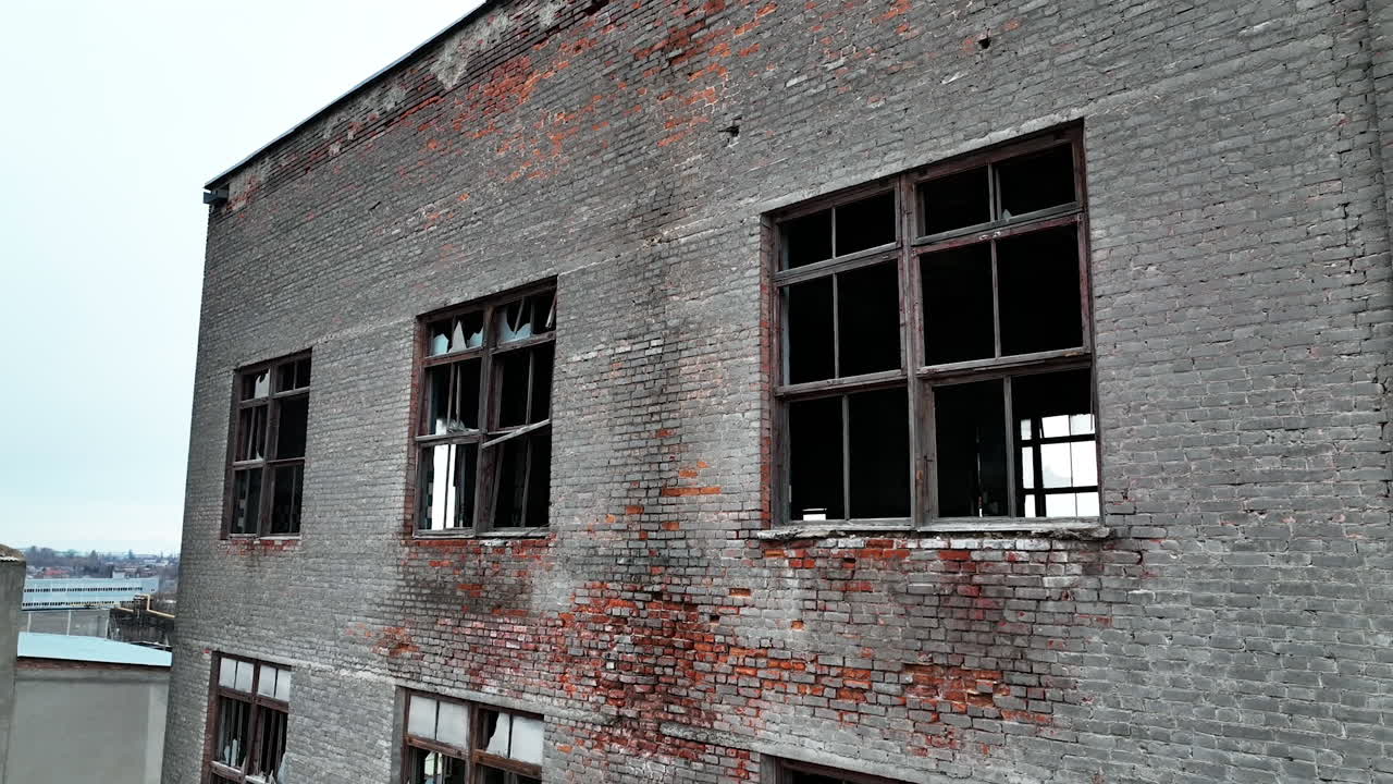 Forlorn building with crashed windows. Approaching to the brick wall of the construction. Industrial chimney pipe at the backdrop.