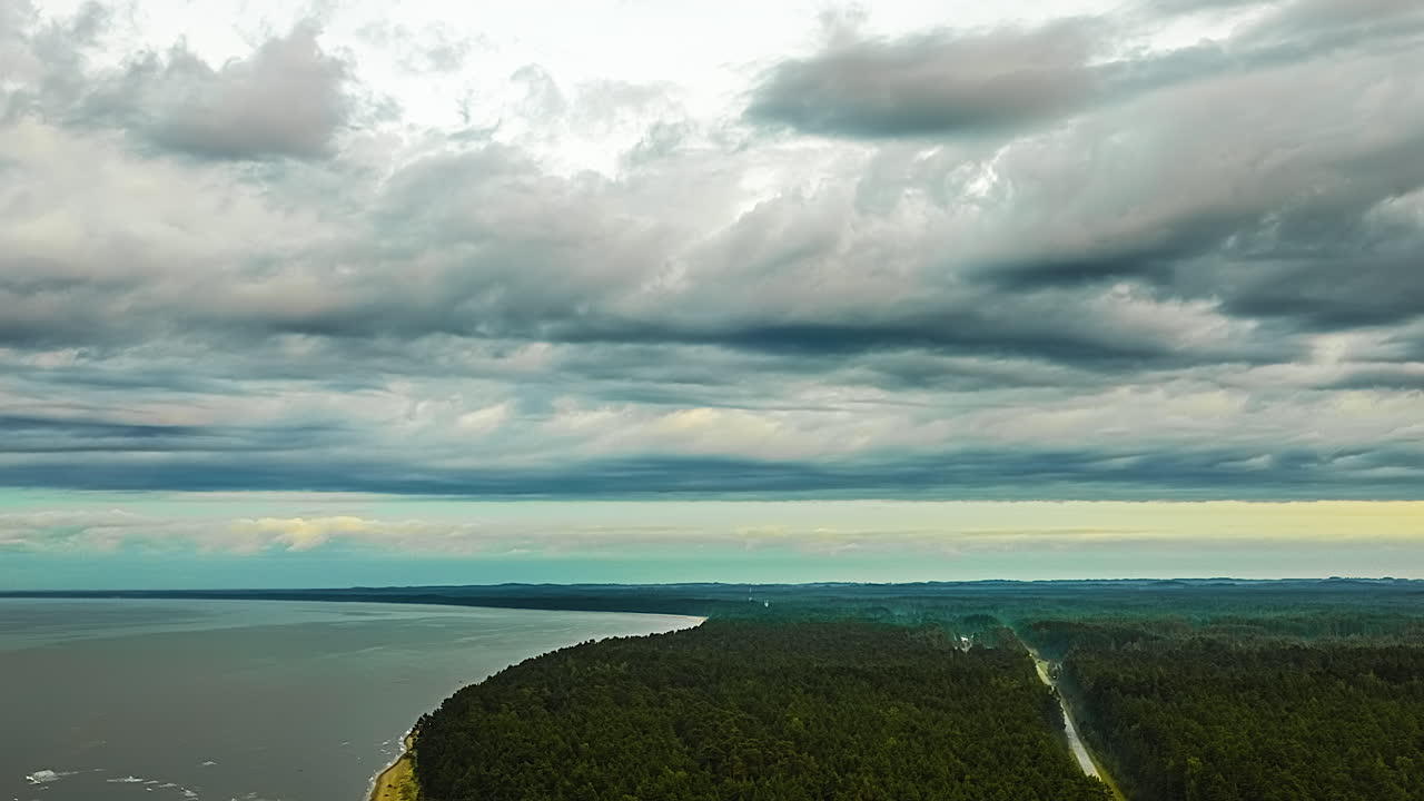 Aerial time lapse shows dramatic clouds moving quickly over a vast green pine forest bordering the Baltic Sea coastline in Latvia with a road running parallel