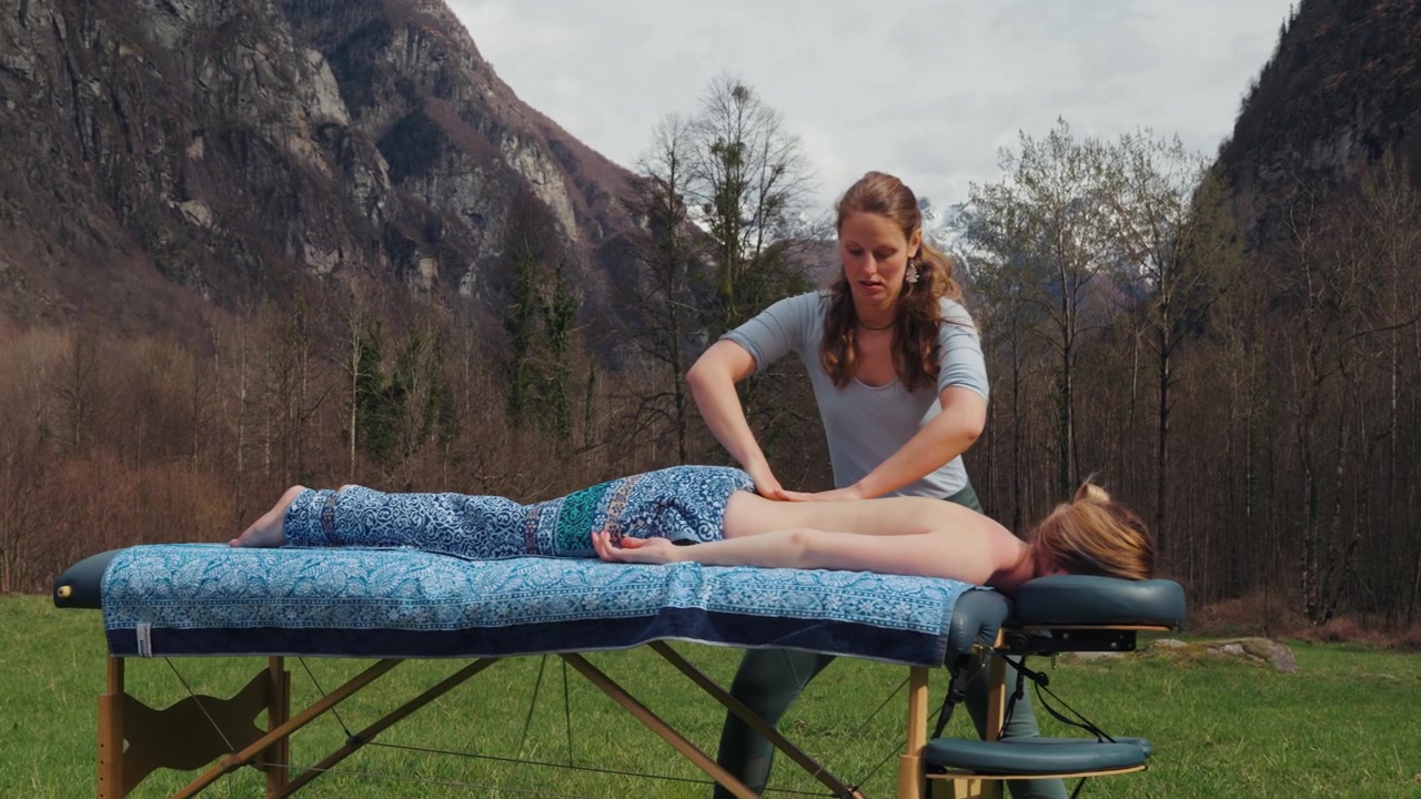 Massageterapeut behandler kvinde på udendørs bord i en alpedal med bjerglandskab, indzoomende skud.
