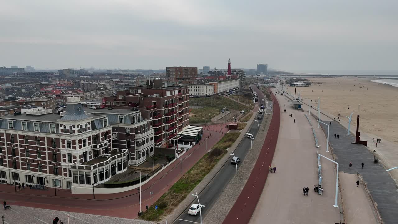 Driving cars and walking people along promenade of Scheveningen Town, Netherlands. Hotel and Apartments with north sea view. Cloudy day with lighthouse in distance. Aerial Forward wide shot.