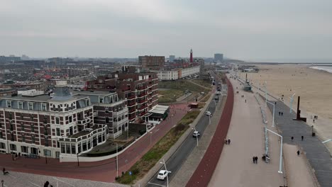 driving cars and walking people along promenade of scheveningen town, netherlands. hotel and apartments with north sea view. cloudy day with lighthouse in distance. aerial forward wide shot.