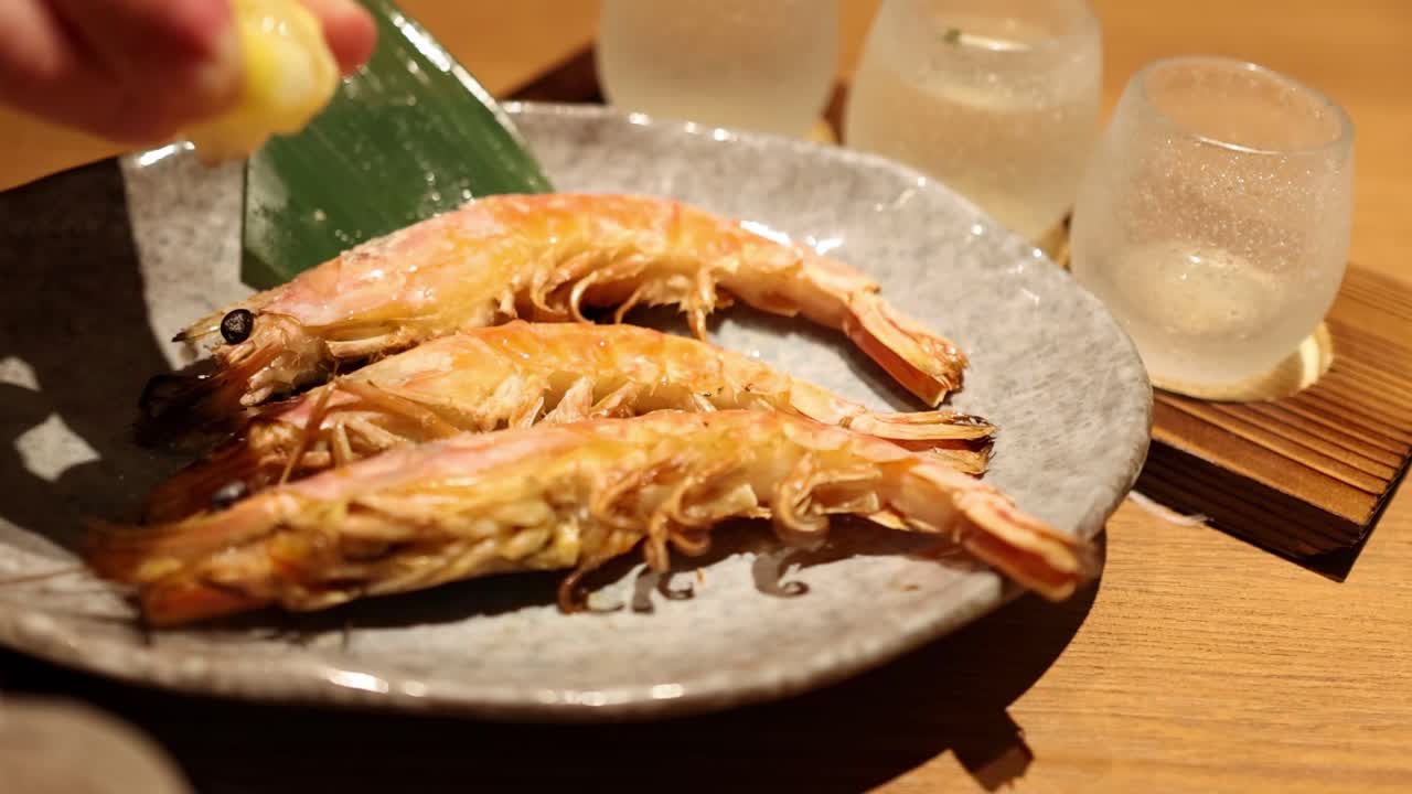 A hand squeezes lemon over grilled shrimp arranged on a plate with a leaf garnish and glasses in the background.