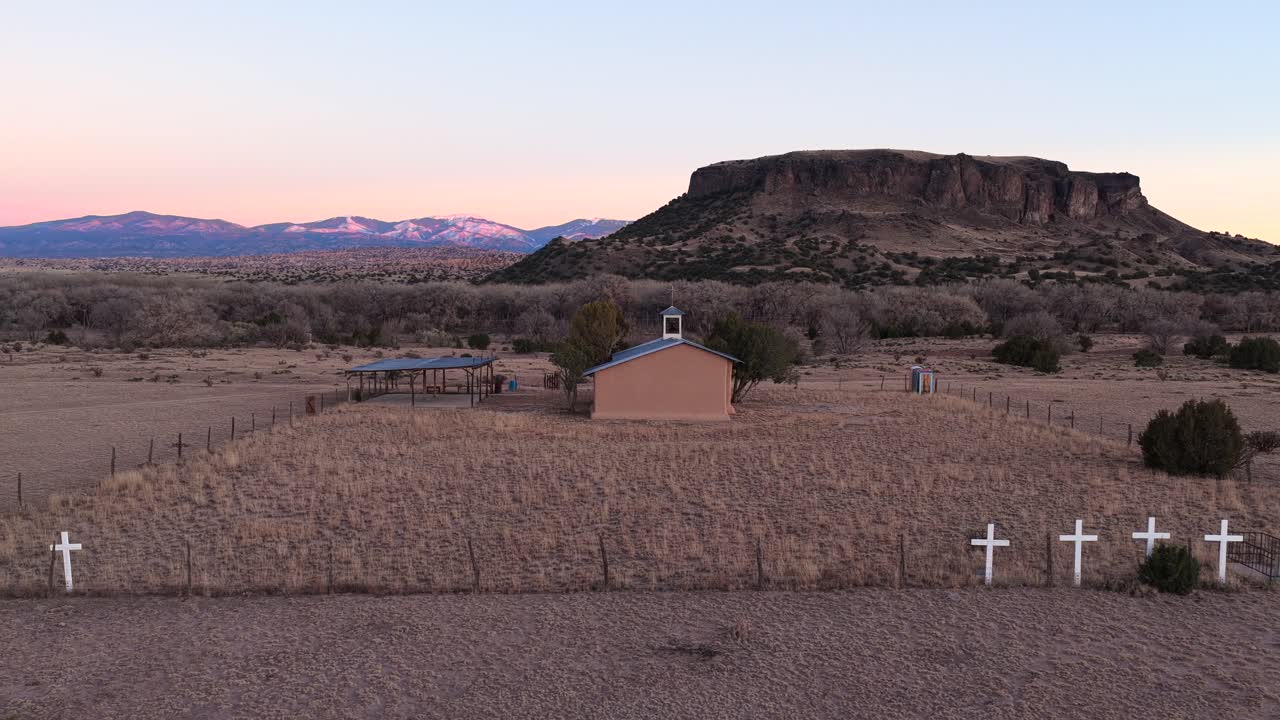 Un drone di alta gamma orbita attorno al campanile di una chiesa rurale al crepuscolo, rivelando delle croci bianche su una recinzione perimetrale. Sullo sfondo, una mesa frastagliata e cime innevate. Perfetto per le aperture di documentari.