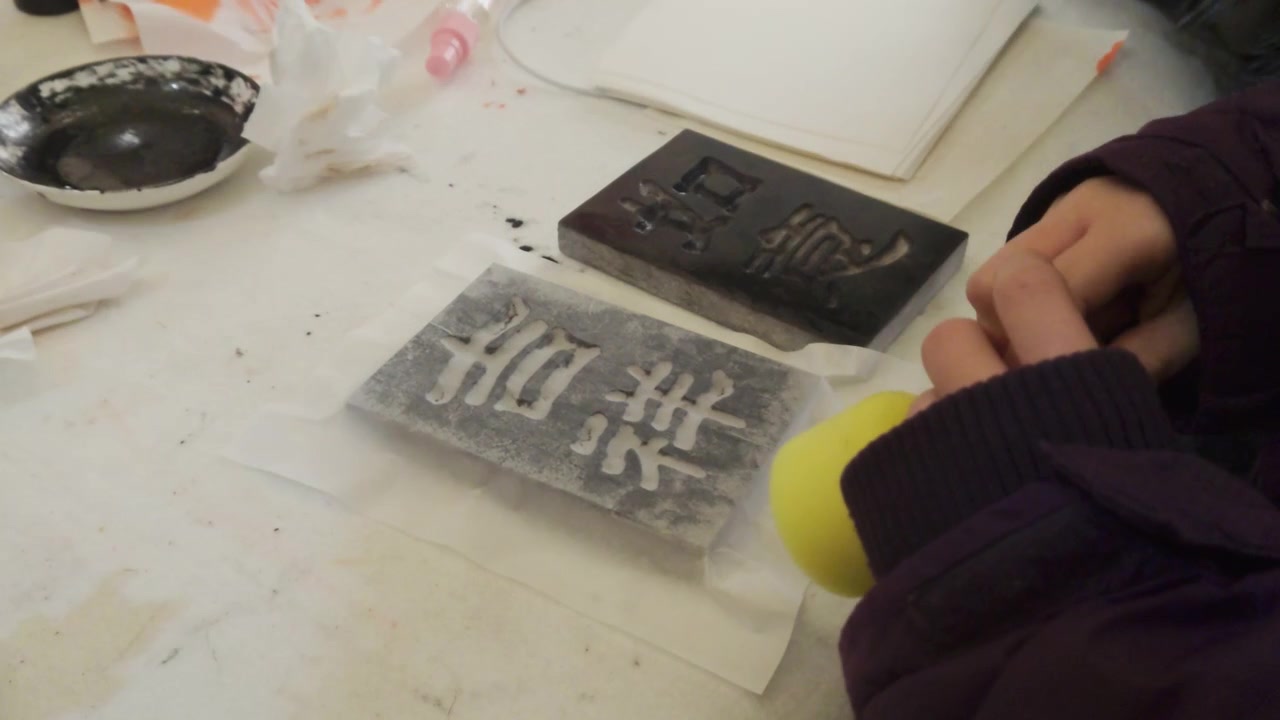 屋外での固定クローズアップショット。伝統的な中国書道の木版印刷デモンストレーションのため、作業台の上で小さな白い皿に墨を準備する講師を捉える。
