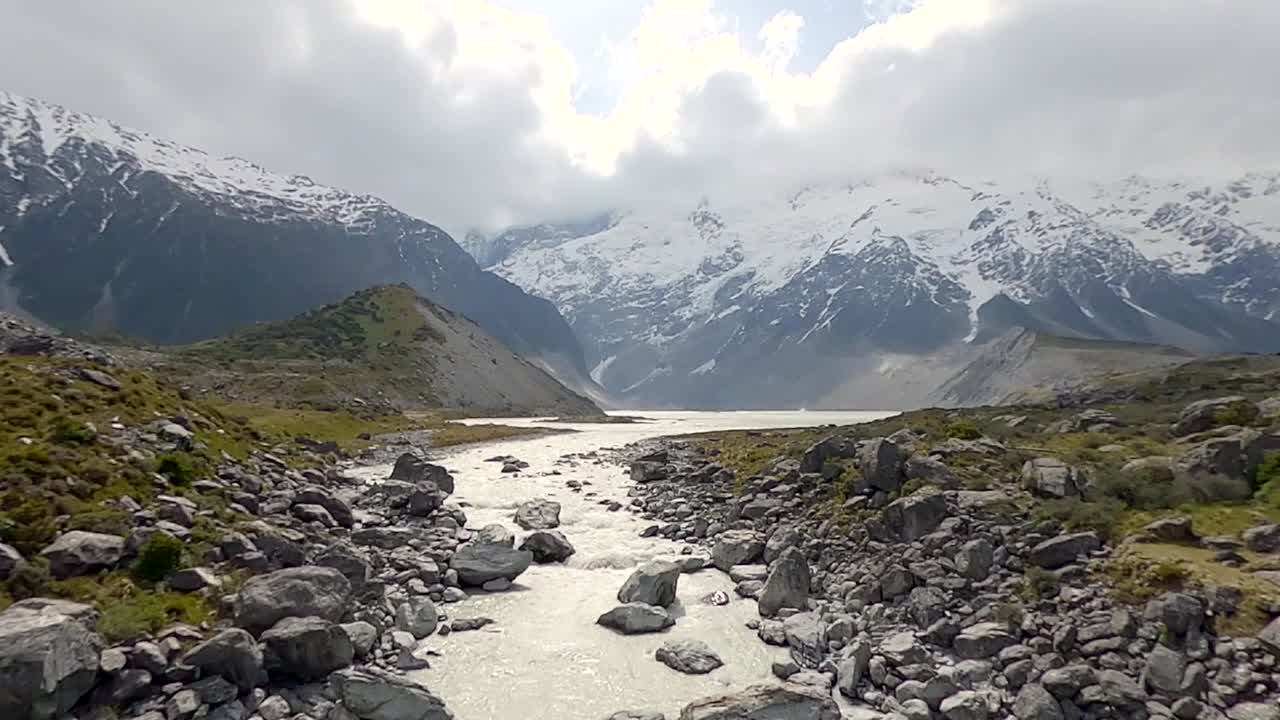Una vista paesaggistica della Hooker Valley in Nuova Zelanda con un fiume che scorre in primo piano e montagne innevate sullo sfondo (slow motion)