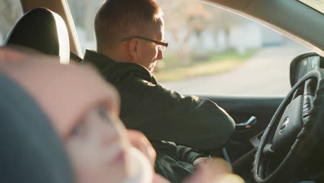 a man in a green jacket is seated in a car, holding a phone in one hand while reaching for his seatbelt. with a blurred view of a young girl in the background