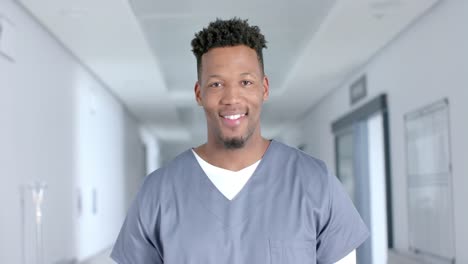 Portrait-of-happy-biracial-male-doctor-wearing-scrubs,-smiling-in-corridor,-slow-motion