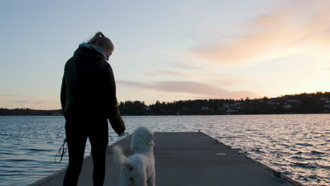 Mujer-Caminando-Con-Caniche-Blanco-Feliz-En-El-Embarcadero-Junto-Al-Agua,-Cámara-Lenta