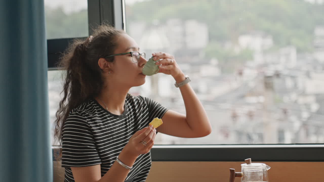 Hóspede de hotel com vista para a cidade comendo biscoito na janela, telhados panorâmicos e horizonte, camisa listrada, óculos, bule na mesa, olhar contemplativo, pausa na viagem, momento calmo de café da manhã no quarto de hotel.