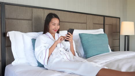 Delighted-asian-lady-scrolling-on-her-phone-on-bed-in-a-bathrobe