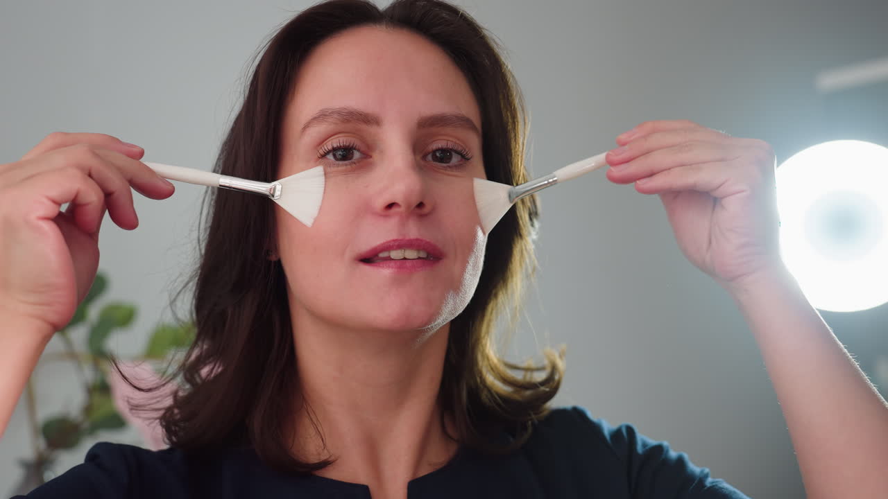 portrait view of cheerful female cosmetologist teasingly brushing cheeks with twin silicone facial brushes under ring light in modern bright spa room with floral decor and skincare products