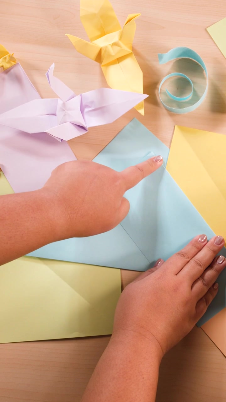Overhead shot of hands carefully folding blue paper on a desk with pastel origami crafts