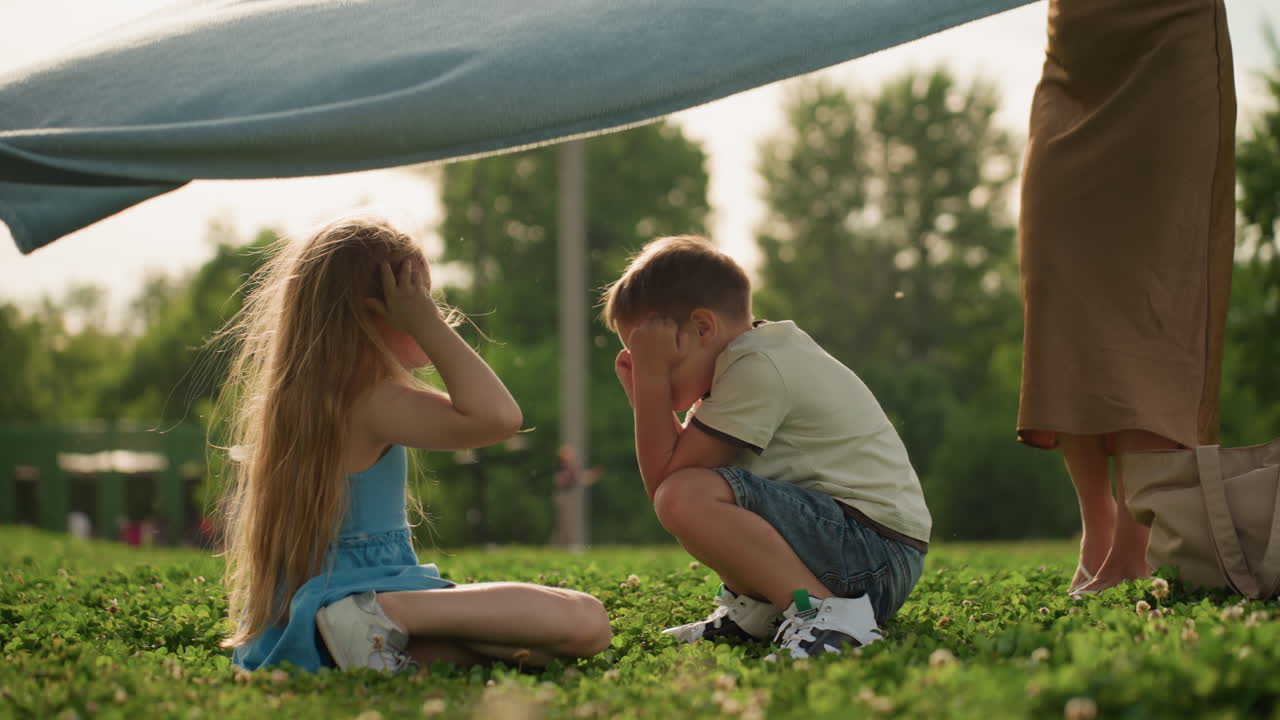 Picnic moms holding two edges of cloth while kids in center laugh and play as fabric swings over them in sunny park, green grass background, casual summer clothes, joyful family bonding moment