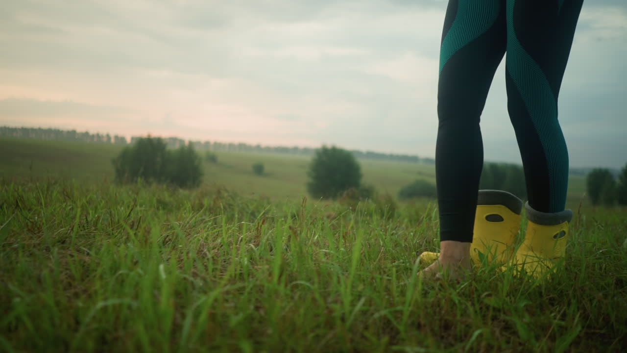 초록색 슈트와 레을 입은 여성은 멀리 나무가 있는 넓은 풀에 서서 노란색 부트를 제거하기 위해 벌거벗은 발을 들어 올립니다.