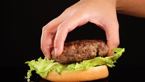 a hand carefully places a cooked beef patty onto iceberg lettuce atop a burger bun, shot in close-up against a black background with studio lighting