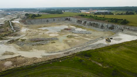 aerial drone view descending and flying lower in front of an yellow colored excavator that is working in a large limestone quarry mine pit by green countryside during a cloudy day, highway visible