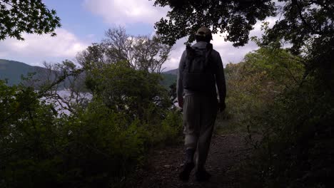 Male-hiker-walking-through-green-forest-next-to-beautiful-lake-and-blue-sky