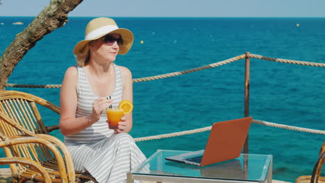 Frauentouristin,-Die-Sich-In-Einem-Café-Mit-Blick-Auf-Das-Meer-Entspannt-Und-In-Der-Nähe-Einen-Cocktail-Trinkt,-Ist-Ein-Laptop-An
