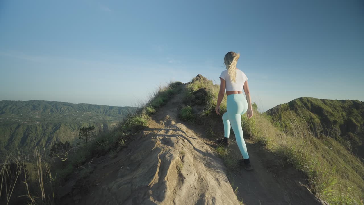mujer rubia caminando por la cresta en el monte batur durante la caminata matutina, alcanzando las manos arriba