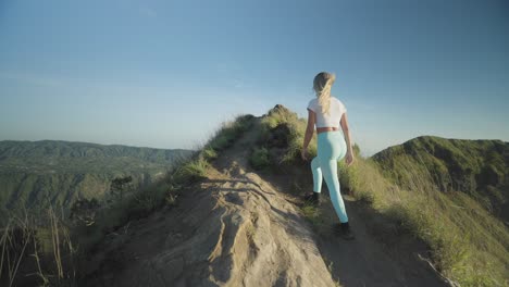 blond woman hiking on ridge on mount batur during morning trek, reaching top hands up