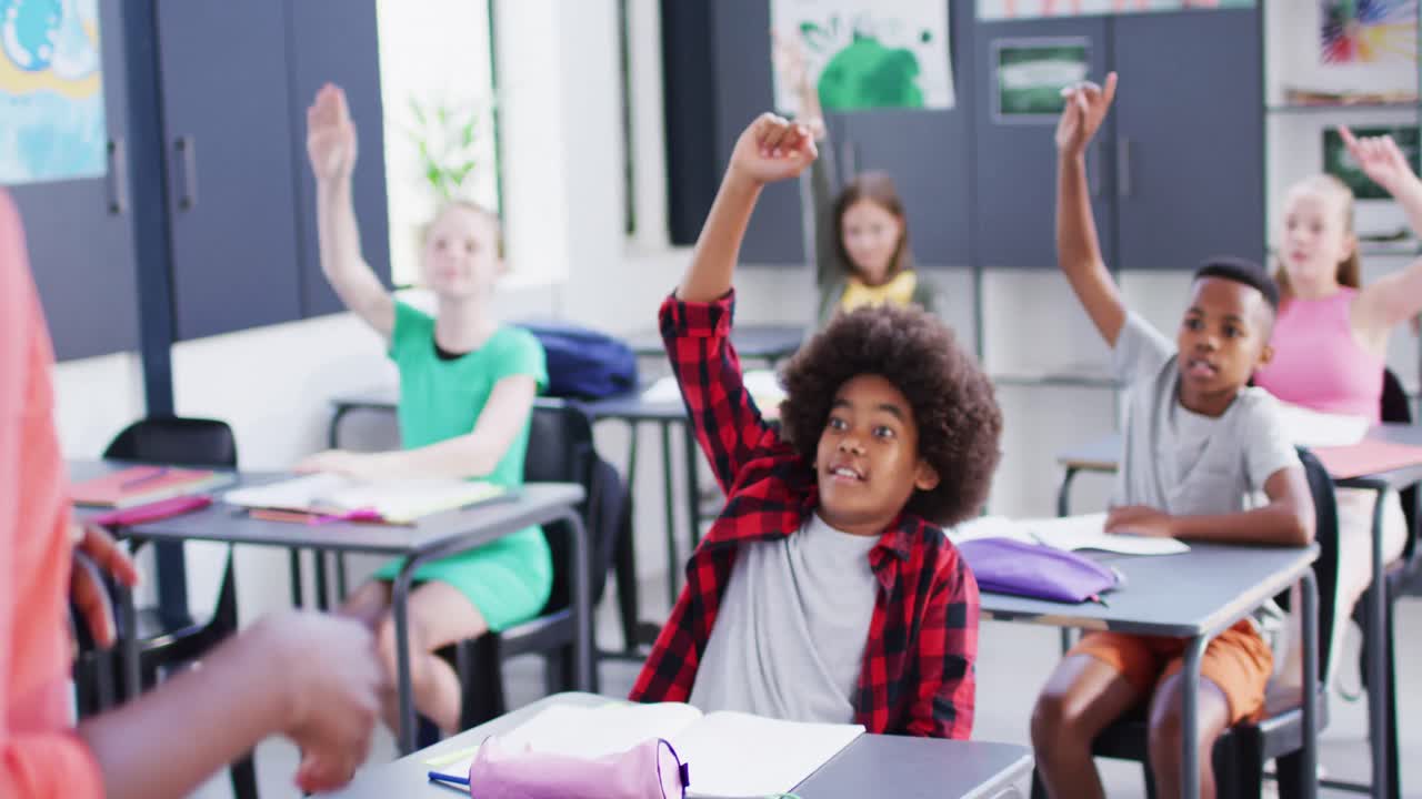 Diverse female teacher and happy schoolchildren at desks raising hands in school classroom