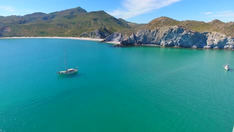 un grupo de yates de vela de casco único anclados en una bahía azul turquesa brillante a lo largo de una playa prístina en la base de pequeñas islas montañosas