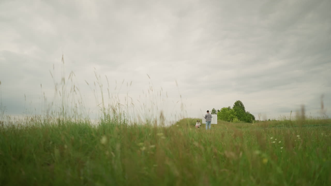 A skilled person wearing a hat and checkered shirt is deeply focused on painting on a board in the middle of a tall grass field under a cloudy sky. Nearby, a woman sits comfortably on a chair